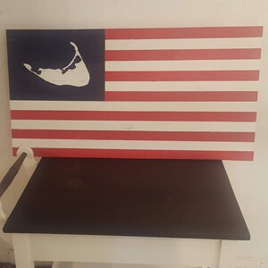 May include: A rectangular wooden sign featuring a painted design of the American flag with red and white horizontal stripes and a blue canton. A white silhouette of Cape Cod is centered within the blue field of the flag on this wall decor piece.