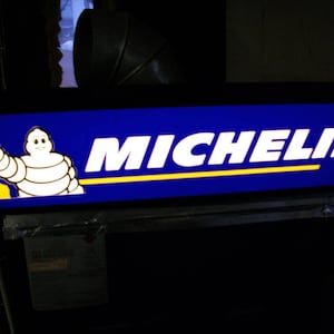 May include: A blue and yellow sign with the Michelin logo and the Michelin Man waving. The sign is lit up and has a black frame.