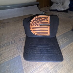 Peut inclure: Casquette de camionneur noire avec un patch en cuir marron représentant le logo Cummins et un motif de drapeau américain.