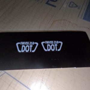 May include: Close-up of a black surface with two white stickers that read "FMVSS 218 DOT" in a white, angled bracket.