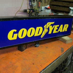 Puede incluir: Un letrero iluminado negro y azul con la palabra "GOODYEAR" en letras amarillas. El letrero tiene un logotipo de pie alado.