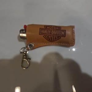 May include: A brown leather lighter case with the Harley-Davidson Cycles logo embossed on the front. The case has a gold-colored metal clip and holds a red and gold lighter.