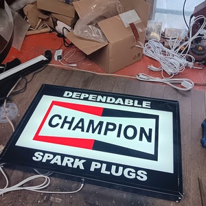 以下が含まれることがあります： 「Dependable Champion Spark Plugs」と書かれた白黒の看板。看板は白い光で照らされています。