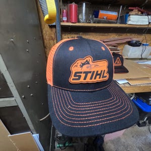 Op de afbeelding: Zwarte en oranje truckerpet met het woord "STIHL" in oranje blokletters. De pet heeft een zwarte klep met oranje stiksels en een oranje mesh achterpaneel. Op de achtergrond is een tweede pet met een oranje "A"-logo zichtbaar.
