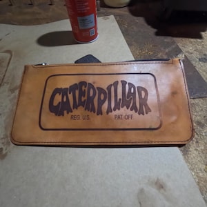 May include: A tan leather pouch with a zipper closure. The pouch features the word "CATERPILLAR" in a brown, stylized font within a rectangular frame. Below the word are the words "REG. U.S." and "PAT. OFF."