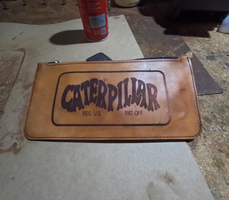 May include: A tan leather pouch with a zipper closure. The pouch features the word "CATERPILLAR" in a brown, stylized font within a rectangular frame. Below the word are the words "REG. U.S." and "PAT. OFF."