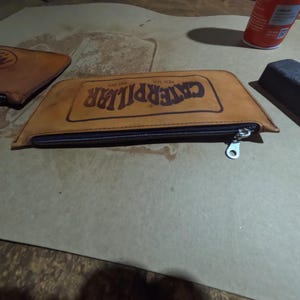 May include: A tan leather pencil case with a black zipper and the word "CATERPILLAR" printed in dark brown. The pencil case is rectangular with a trapezoid shape. A second leather item is visible in the background.