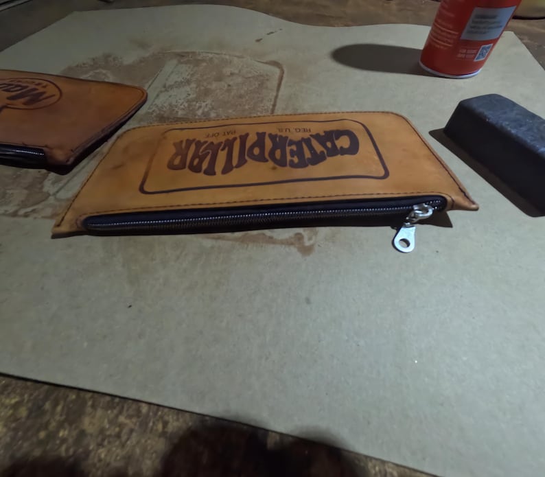 May include: A tan leather pencil case with a black zipper and the word "CATERPILLAR" printed in dark brown. The pencil case is rectangular with a trapezoid shape. A second leather item is visible in the background.