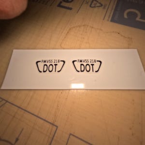 May include: Two white stickers with black text and a black border. Each sticker has the text "FMVSS 218" above the word "DOT" in a stylized font. The stickers are on a white rectangular background.