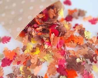 May include: A tan paper bag with white polka dots spilling out small, die-cut paper leaves in shades of red, orange, yellow, and brown. The confetti pieces are shaped like various autumn foliage and are scattered across the surface.