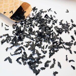 May include: A tan paper bag with a white quatrefoil pattern spilling out a large quantity of small, black, bat-shaped paper confetti pieces scattered across a plain white surface, creating a dark, high-contrast decorative display.