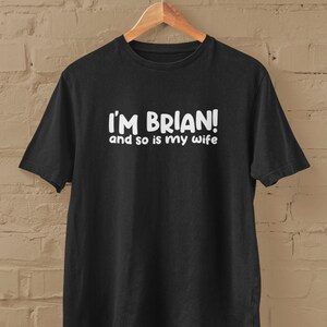 Op de afbeelding: Zwart T-shirt met witte tekst die "Ik ben Brian! en mijn vrouw ook" luidt