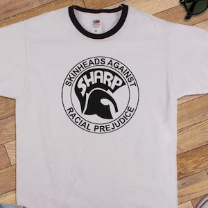 Op de afbeelding: Wit T-shirt met een zwart-witte cirkelvormige afbeelding met de tekst "Skinheads Against SHARP Racial Prejudice". Het shirt heeft een zwart-witte boord.