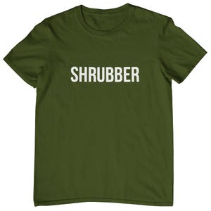 May include: An olive green t-shirt featuring the word "SHRUBBER" in white, bold capital letters. The shirt has a crew neck and short sleeves. The fabric appears soft and comfortable.