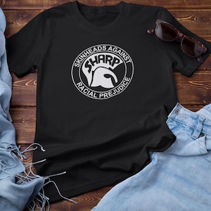 Op de afbeelding: Zwart T-shirt met een witte cirkelvormige afbeelding met de tekst "Skinheads Against SHARP Racial Prejudice". De afbeelding toont een witte Spartaanse helm.