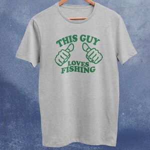 May include: A gray t-shirt with green text that reads "This Guy Loves Fishing". The text is surrounded by two green thumbs pointing to the left and right.