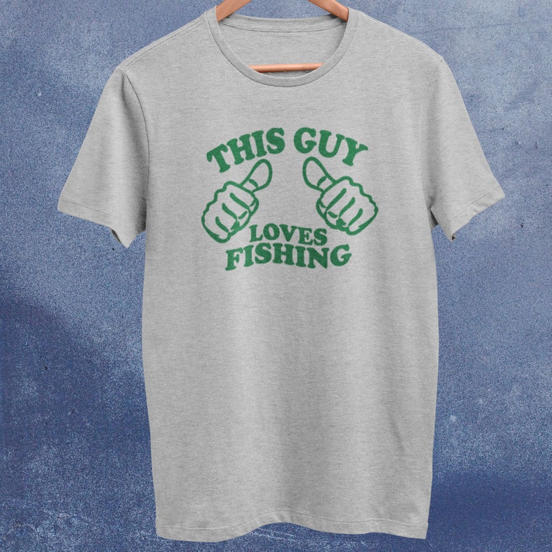 May include: A gray t-shirt with green text that reads "This Guy Loves Fishing". The text is surrounded by two green thumbs pointing to the left and right.