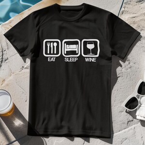 Può includere: T-shirt nero con un design grafico bianco con le parole "EAT SLEEP WINE" con icone corrispondenti di forchetta e coltello, letto e bicchiere di vino. Occhiali da sole e un bicchiere di birra sono visibili sullo sfondo.