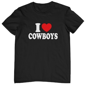 Może przedstawiać: Czarny t-shirt z krótkim rękawem z białym napisem I, czerwonym symbolem serca oraz słowem COWBOYS zapisanym białymi wielkimi literami na środku klatki piersiowej. Koszulka posiada okrągły dekolt oraz standardowy krój.