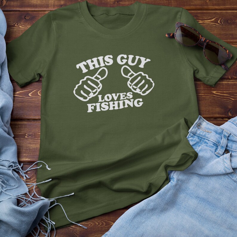 May include: A green t-shirt with white text that reads "This Guy Loves Fishing". The text is surrounded by two white thumbs up symbols.