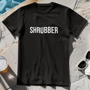 Peut inclure: T-shirt noir avec le mot "SHRUBBER" imprimé en blanc. Le t-shirt est à manches courtes et a un col rond. Des lunettes de soleil et un verre de bière sont visibles en arrière-plan.