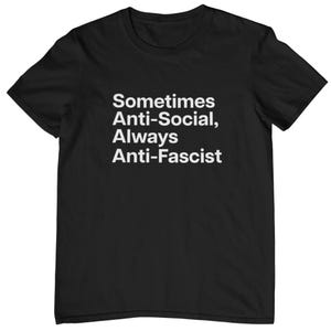 Może przedstawiać: Czarny t-shirt z krótkim rękawem z białym napisem na piersi: Sometimes Anti-Social, Always Anti-Fascist. Odzież posiada okrągły dekolt i standardowy krój, podkreślający kontrast między ciemną tkaniną a pogrubionymi literami.