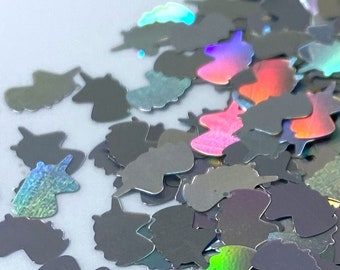 May include: A pile of iridescent silver glitter shaped like unicorns. The glitter is scattered on a white surface.