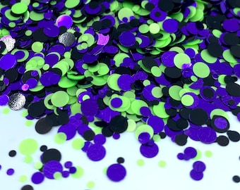 May include: A pile of green, purple, and black glitter confetti. The confetti is in the shape of circles of various sizes.