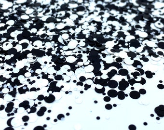 May include: Close-up of a scattering of black and white round sequins. The sequins vary in size and are scattered across a white surface, creating a textured, abstract pattern. The black sequins are solid, while the white ones have a reflective sheen.
