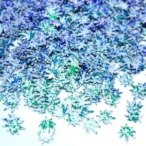 May include: A scattering of silver glitter leaf shapes with blue and green paint splatter details. The glitter is on a white background.
