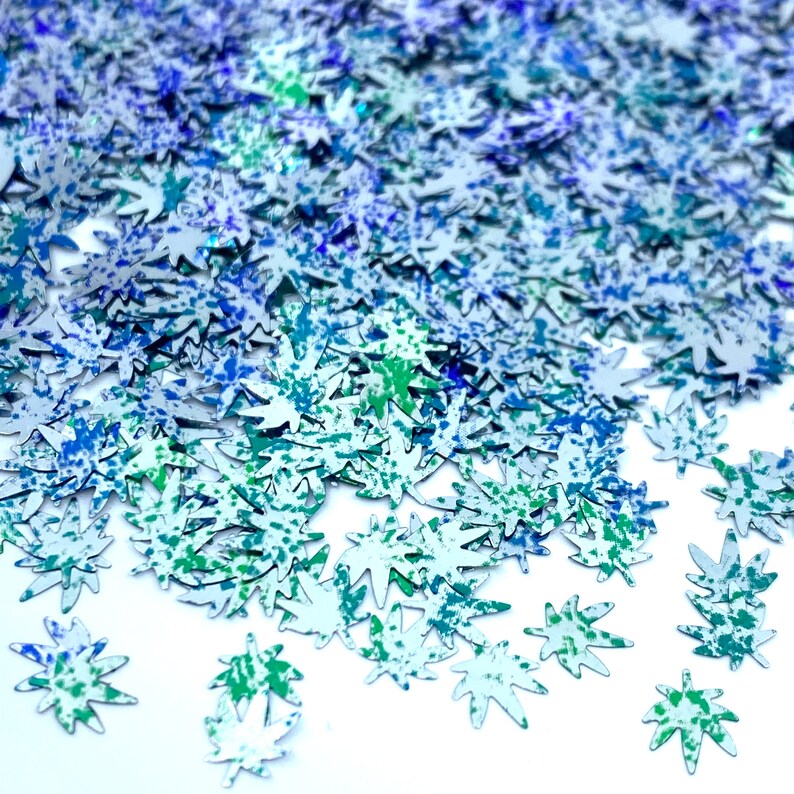 May include: A scattering of silver glitter leaf shapes with blue and green paint splatter details. The glitter is on a white background.