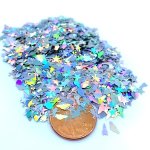 May include: A pile of iridescent holographic glitter in various shapes, including stars, butterflies, and other abstract shapes. A penny is included for scale.