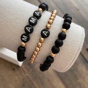 May include: Three beaded bracelets. Two black bracelets with gold accents and the word "MAMA" spelled out in white letters. The third bracelet is gold with a single black letter "A".
