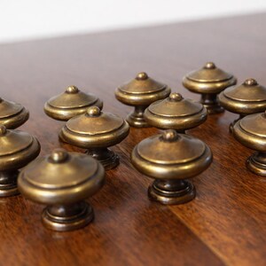 May include: A collection of antique brass cabinet knobs. Each knob features a rounded top and a decorative base. The knobs are arranged on a dark wood surface, showcasing their vintage design and warm, metallic color.