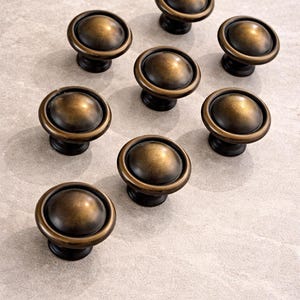 May include: Nine antique bronze cabinet knobs arranged on a light gray surface. Each knob has a rounded, domed top and a circular base. The knobs are made of metal and have a warm, aged finish, suggesting a vintage or classic style.