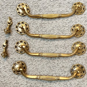 May include: Four gold-toned cabinet pulls with ornate, decorative details. Each pull has a rounded, decorative knob and a long, curved handle with a ribbed design.