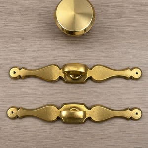 May include: A set of brass hardware consisting of one circular knob with a smooth, polished finish and two elongated, decorative drawer pulls. Each pull features a central ring handle and two screw holes at the ends for mounting purposes.