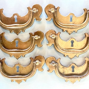 May include: Six antique gold-toned cabinet drawer pulls with a keyhole design. Each pull has a decorative, ornate, scalloped edge.