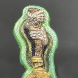 May include: A green and gold broom with a hand holding the handle. The broom is made of a soft, textured material and has a gold handle with a brown base.