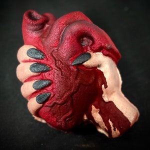 May include: A red heart-shaped object with a textured surface, covered in a dark red substance that appears to be dripping. The object is held in a hand with black fingernails.