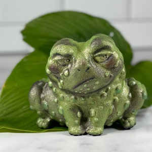 May include: A green ceramic frog figurine with a textured surface and a grumpy expression. The frog is sitting on a green leaf.