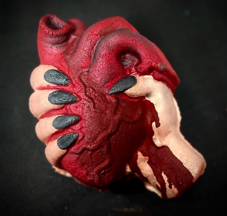 May include: A red heart-shaped object with a textured surface, covered in a dark red substance that appears to be dripping. The object is held in a hand with black fingernails.