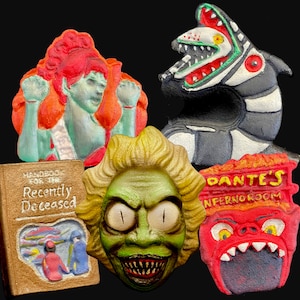 May include: Five colorful, hand-painted figurines, including a green monster with a toothy grin, a red monster with a large mouth, a black and white striped monster with a large mouth, a red-haired woman with a green dress, and a book with the title "Handbook for the Recently Deceased".