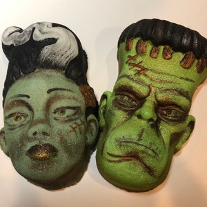 May include: Two green bath bombs shaped like Frankenstein's monster and his bride. The bride has black hair and a white streak in her hair. The monster has black hair and stitches on his forehead. Both have gold accents.