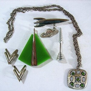 May include: A set of silver tone space-themed jewelry, including a necklace with a square pendant with green stones, a rocket ship brooch, a green glass triangle pendant, a pair of earrings, and a silver tone rocket ship charm.
