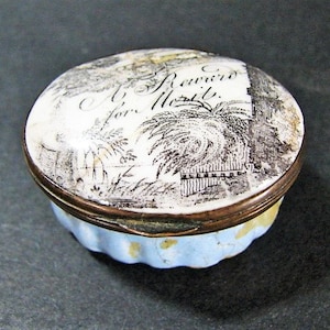 May include: An antique, oval-shaped trinket box with a white lid featuring a black and white landscape illustration and the text "Souvenir A. Mort". The box has a brass rim and a light blue, scalloped base. A vintage decorative item.