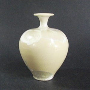 May include: A small, off-white ceramic vase with a rounded body and a narrow neck. The vase has a textured surface and a slightly flared rim. The base of the vase has a small chip. The vase is likely for decorative purposes.