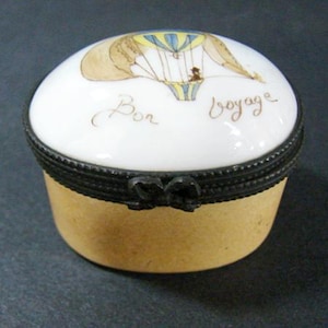 May include: A white porcelain trinket box with a black trim and a black bow. The lid features a hand-painted image of a hot air balloon with the words "Bon Voyage" in brown script.