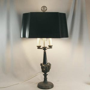 May include: An antique table lamp with a black lampshade and a decorative base. The lamp has three candle-style bulbs and a dark, ornate metal stand. The lamp's base is approximately 8 inches in diameter.