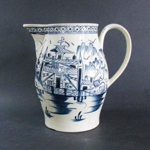 May include: A white ceramic pitcher with a blue and white landscape design. The pitcher features a curved handle and a spout. The design includes buildings, trees, and water, with a decorative border around the rim.